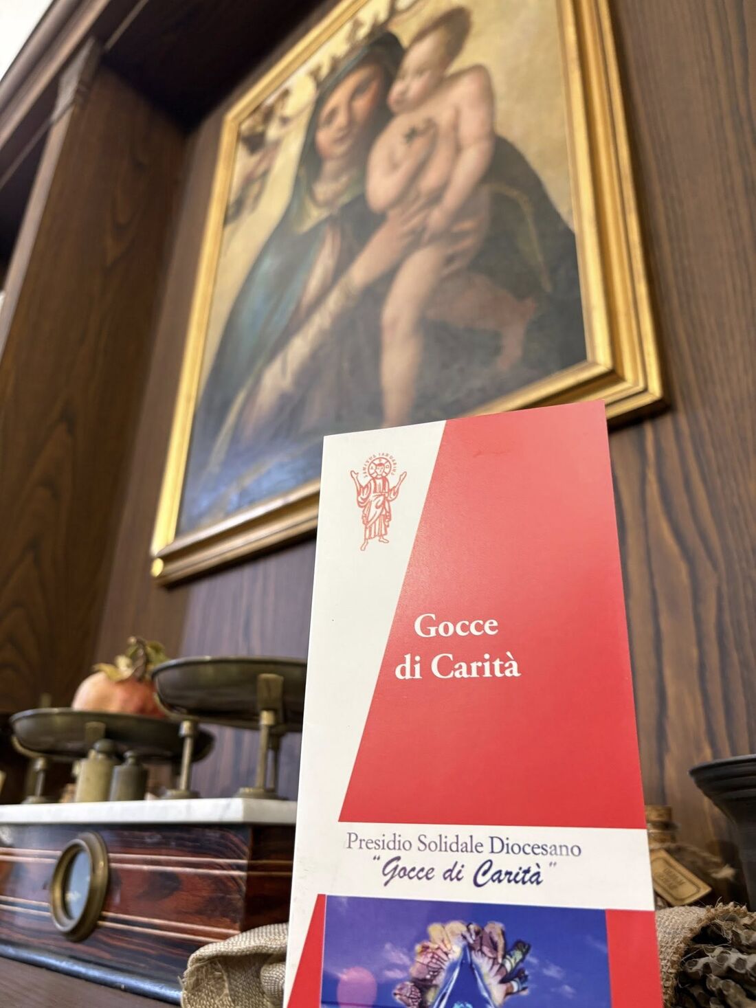 La brochure rossa di Gocce di Carità davanti a un dipinto della Madonna col Bambino